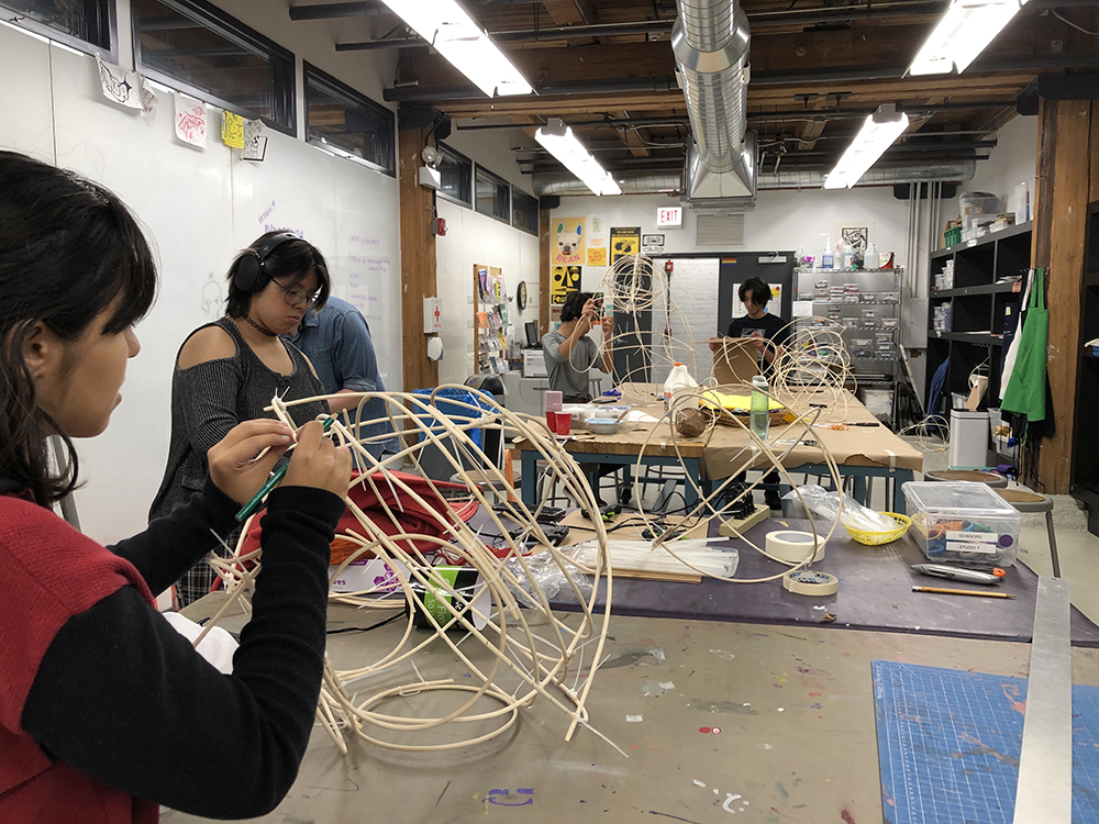 Young artists in the studio use bendable wooden rods to create 3D frames for wearable sculptures.
