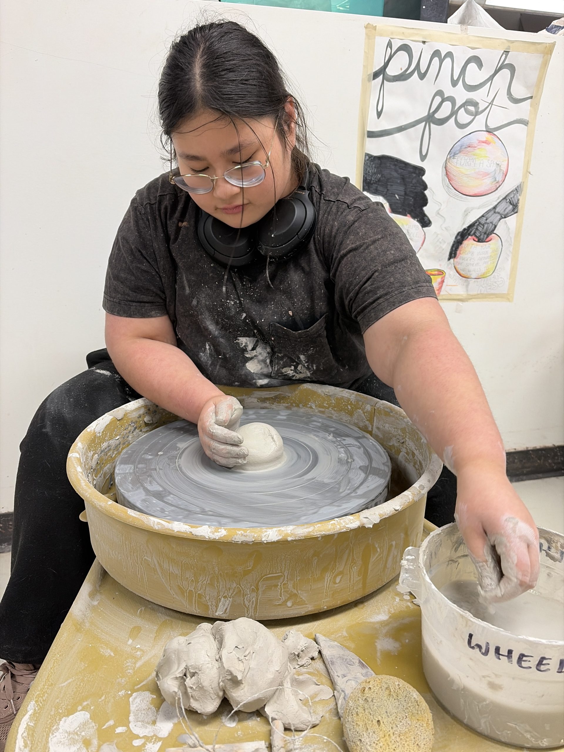 Person wearing glasses and a necklace of headphones shapes gray clay on a spinning pottery wheel.