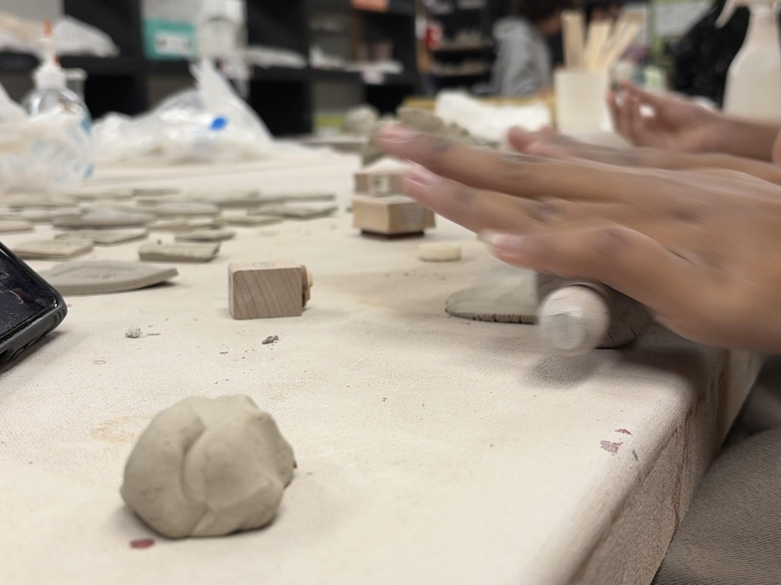People hands shaping clay on a table in a classroom; small clay pieces and wooden blocks scattered around.