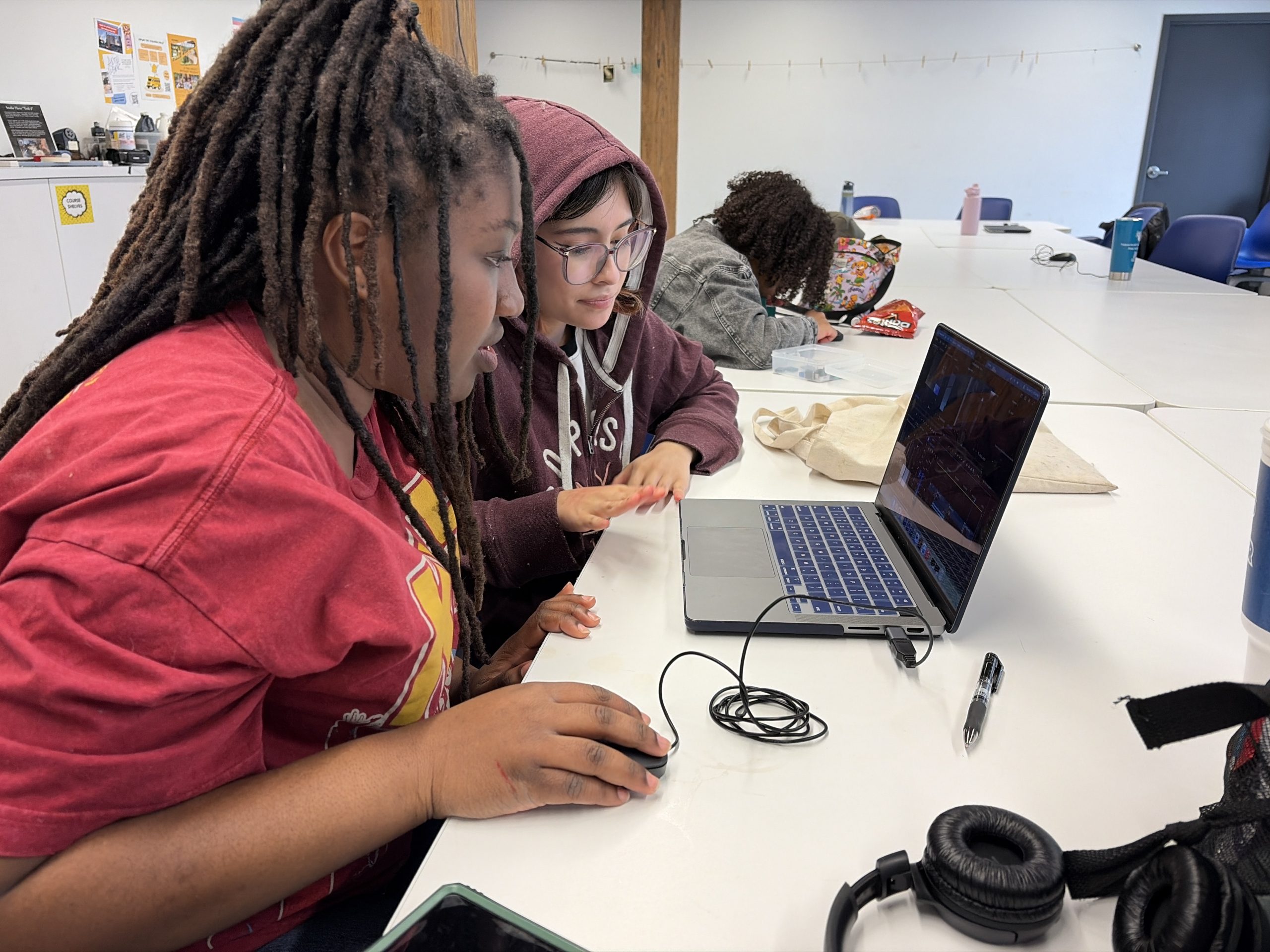 Two students sit at a long white table working with video editing software