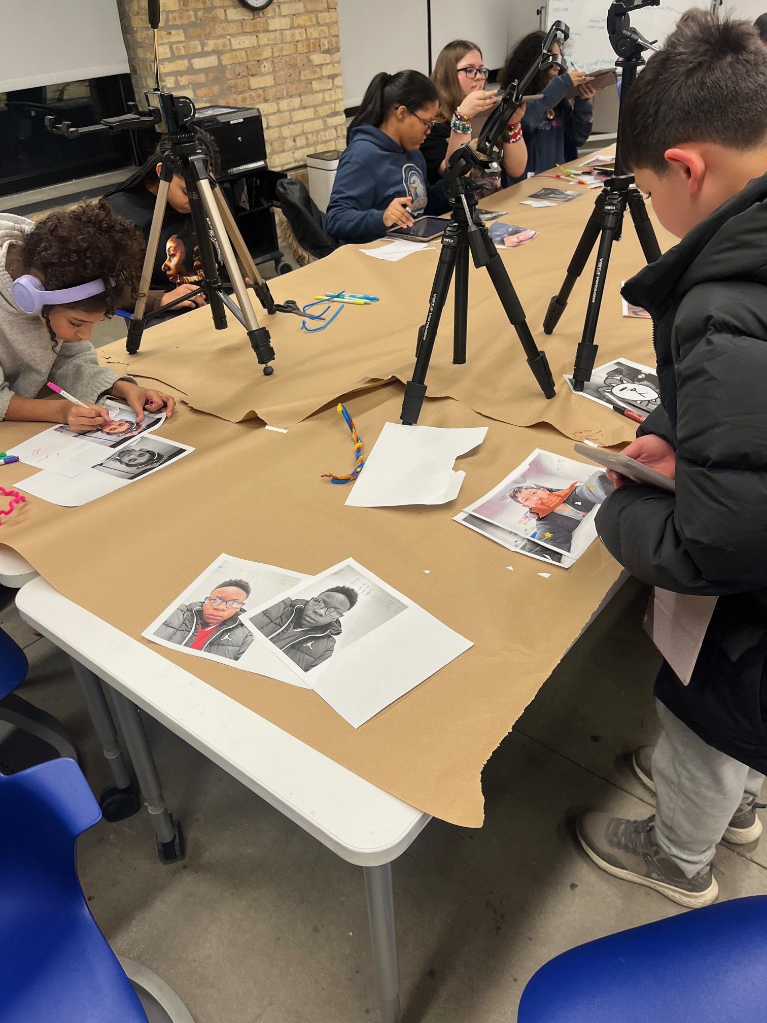 Students work around a table covered in brown paper, with cameras on tripods and printed photos for a photography workshop.