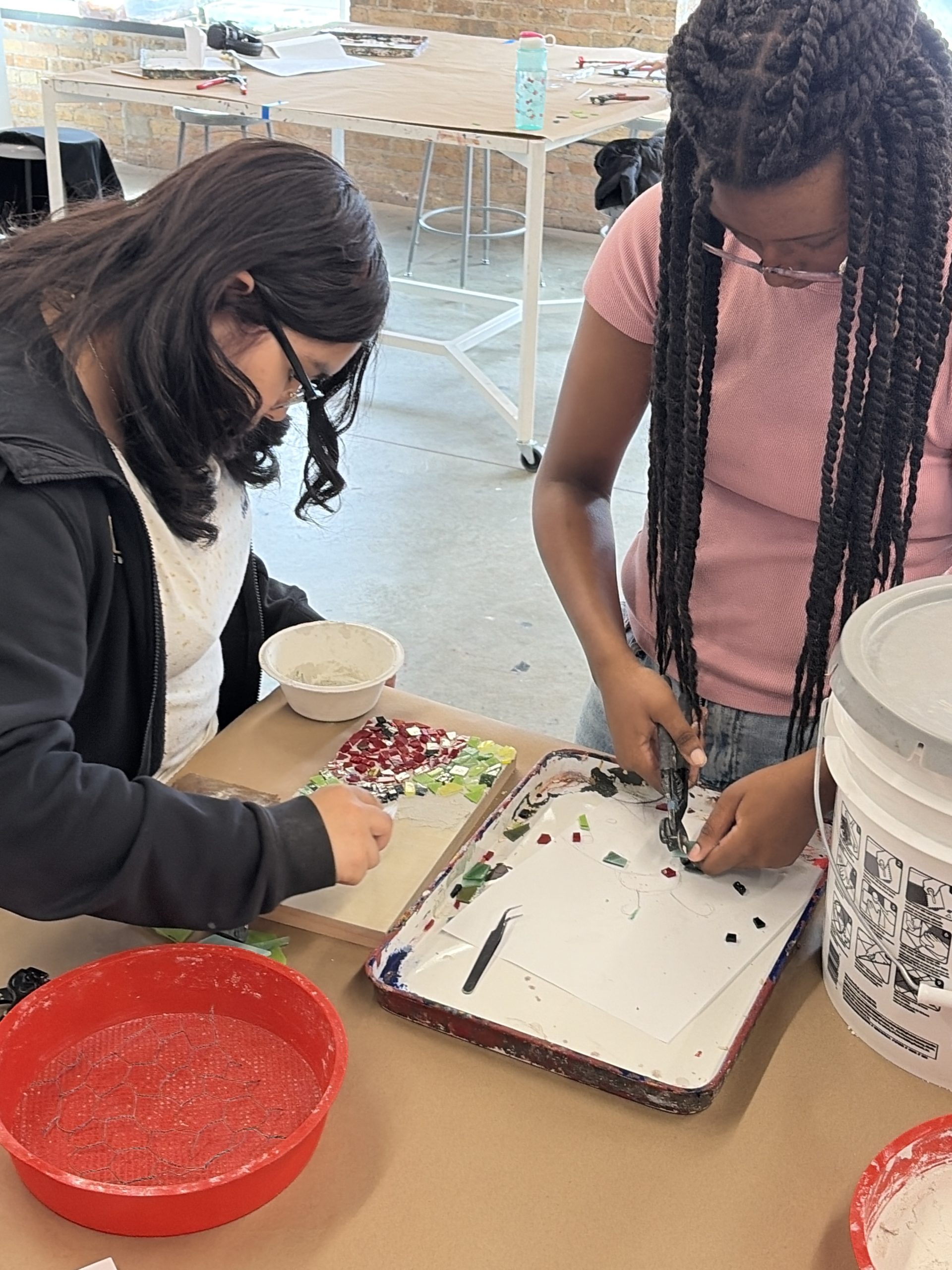 Two young students work on a mosaic art project at a table, placing colorful tile pieces on a tray outdoors.