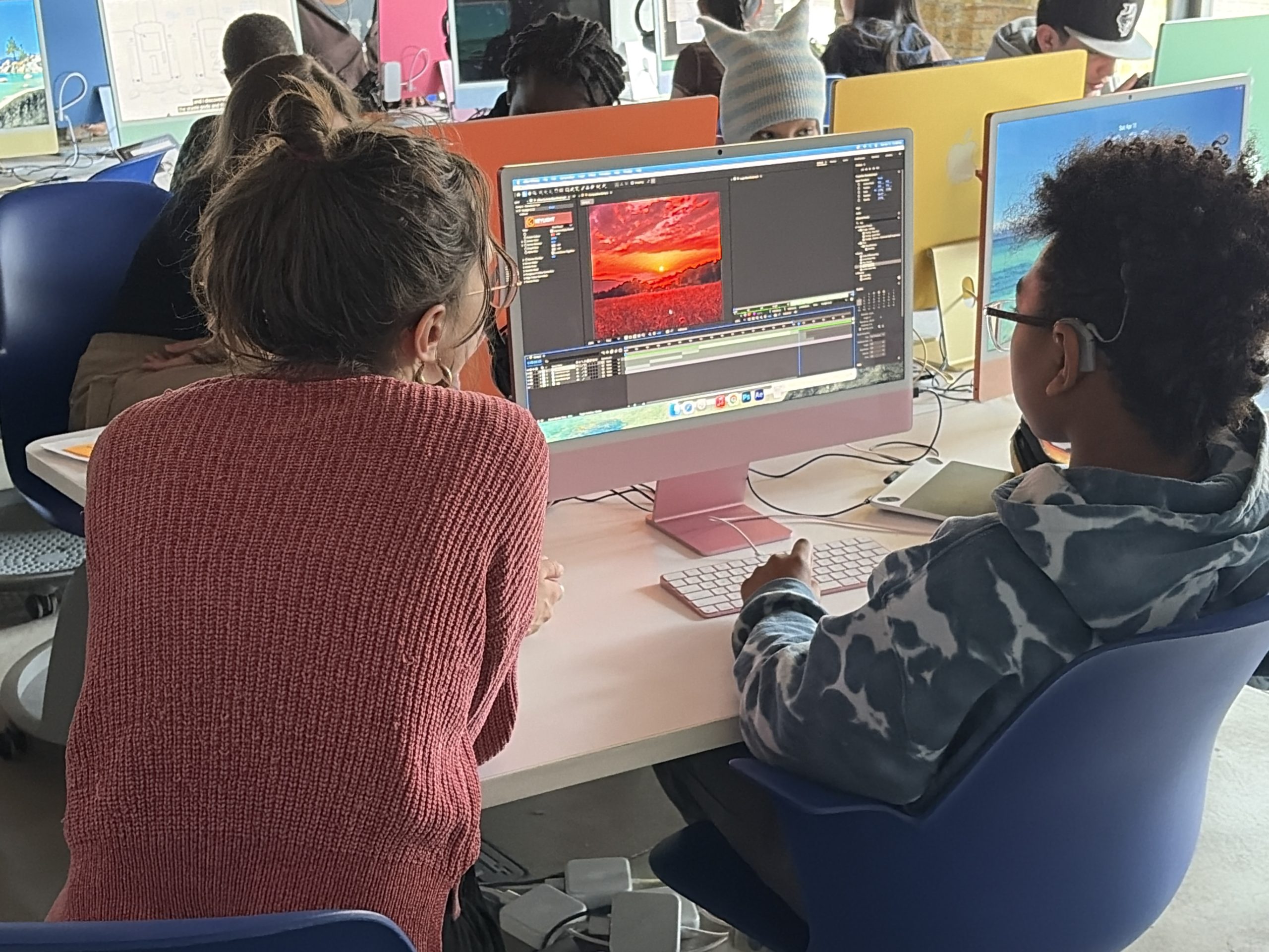 Two students sit at a pink desk editing video on an iMac, monitor showing editing software in a bright classroom.