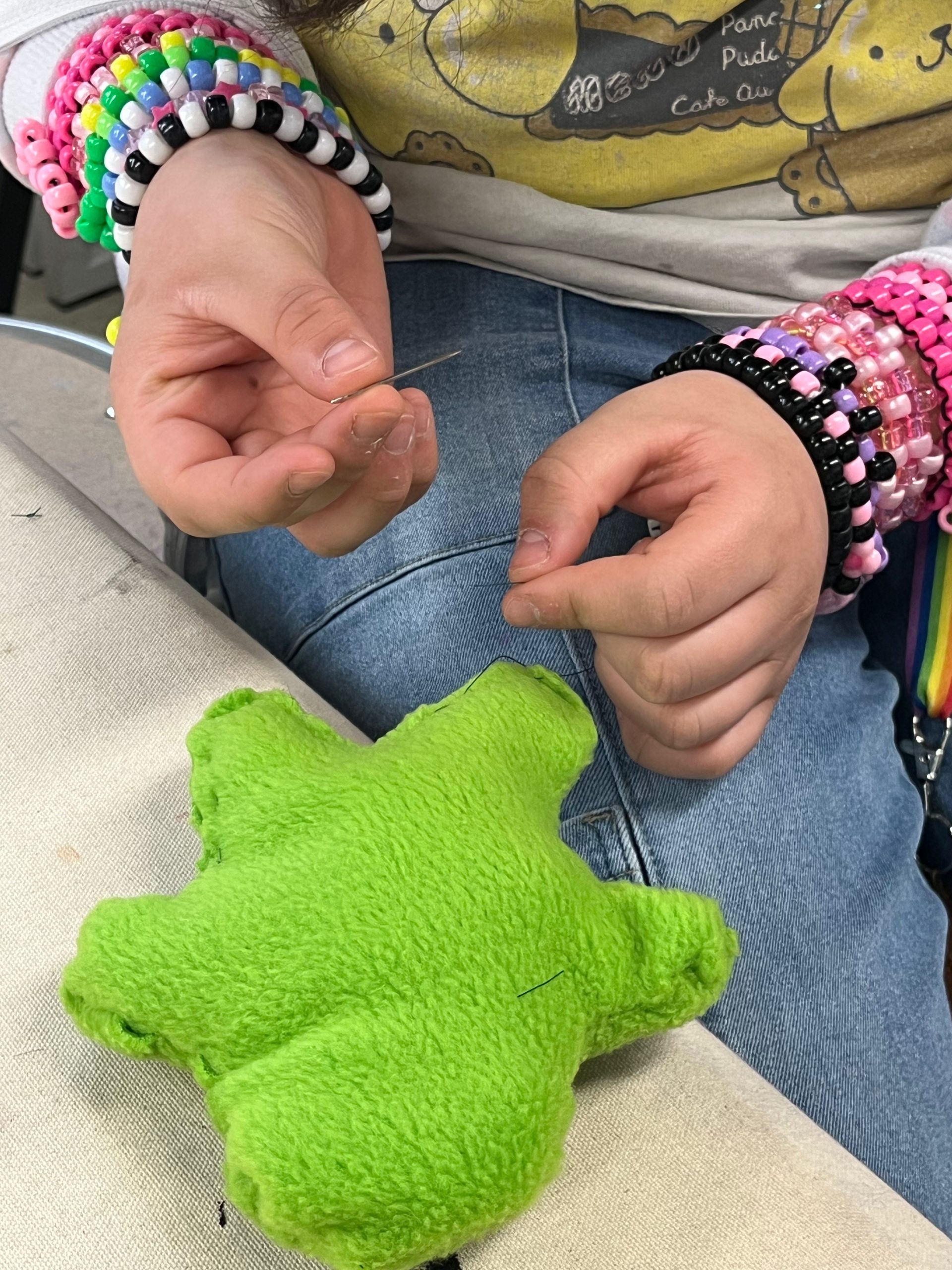 Child's hands threading a needle for crafts; wearing colorful beaded bracelets, pinks and greens, with a bright green star plush nearby on fabric.