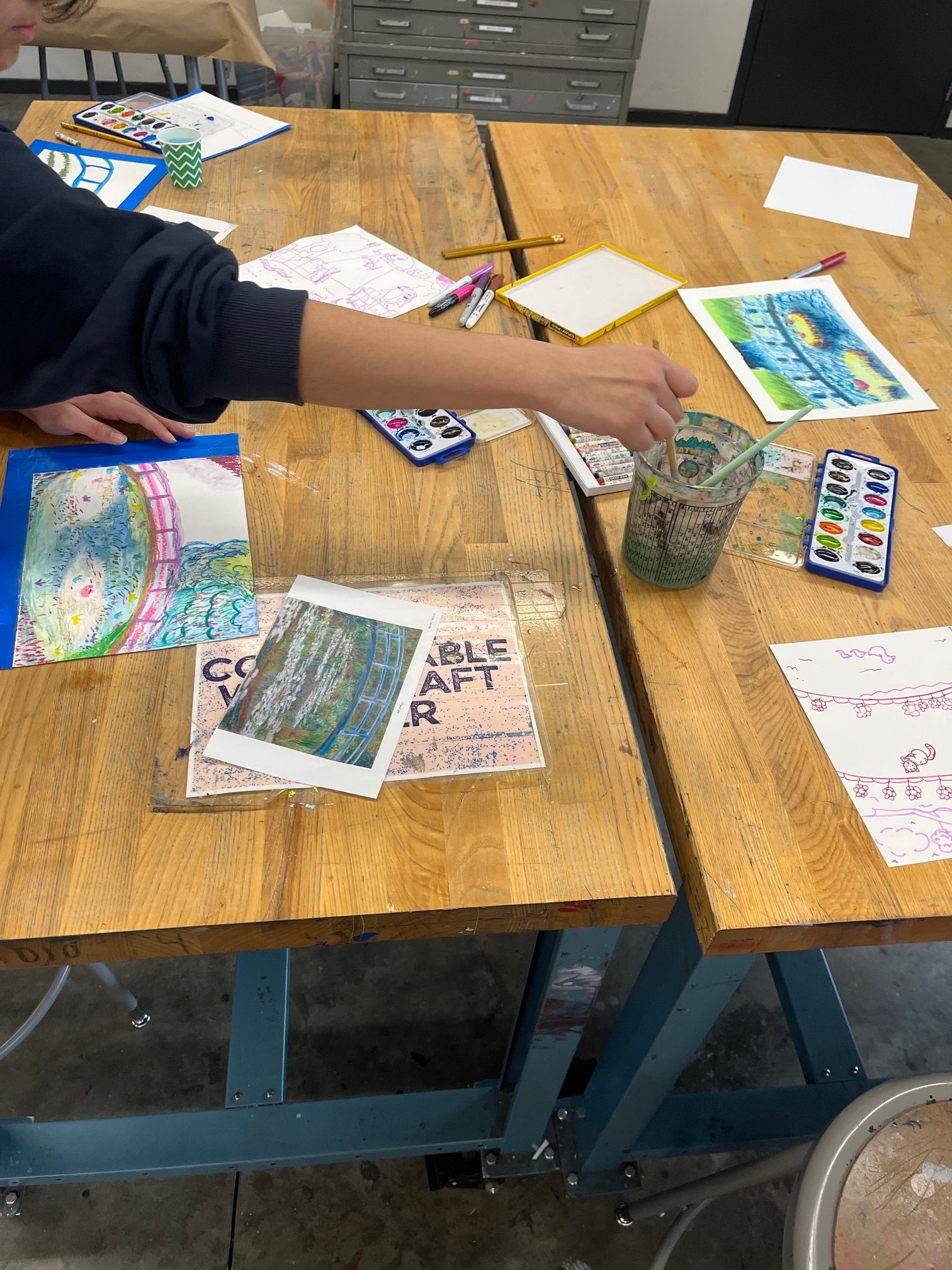 Student reaches into a cup of water while painting at a cluttered wooden art table, with watercolor paints, brushes, and several colorful drawings scattered around.