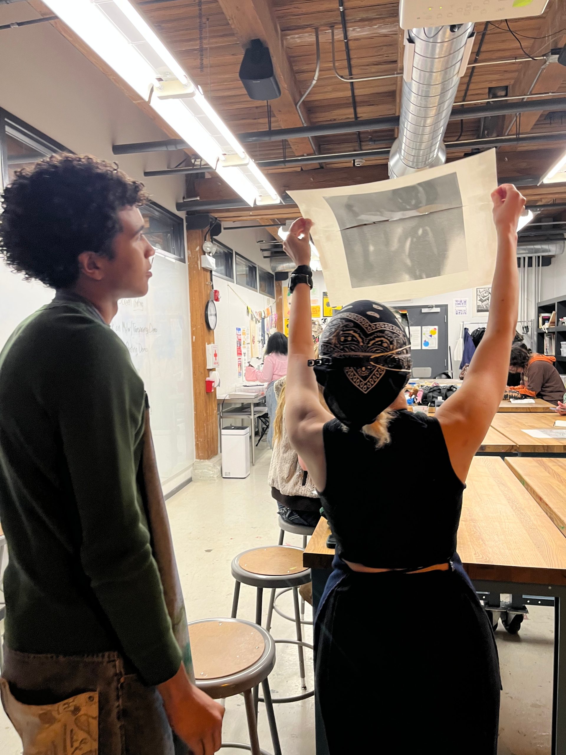 A person with a bandana raises a large printed artwork above their head in a busy classroom studio while another student watches nearby.