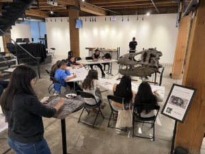 Group of students sketching at long tables in a museum studio, with a large dinosaur skull model on display nearby.