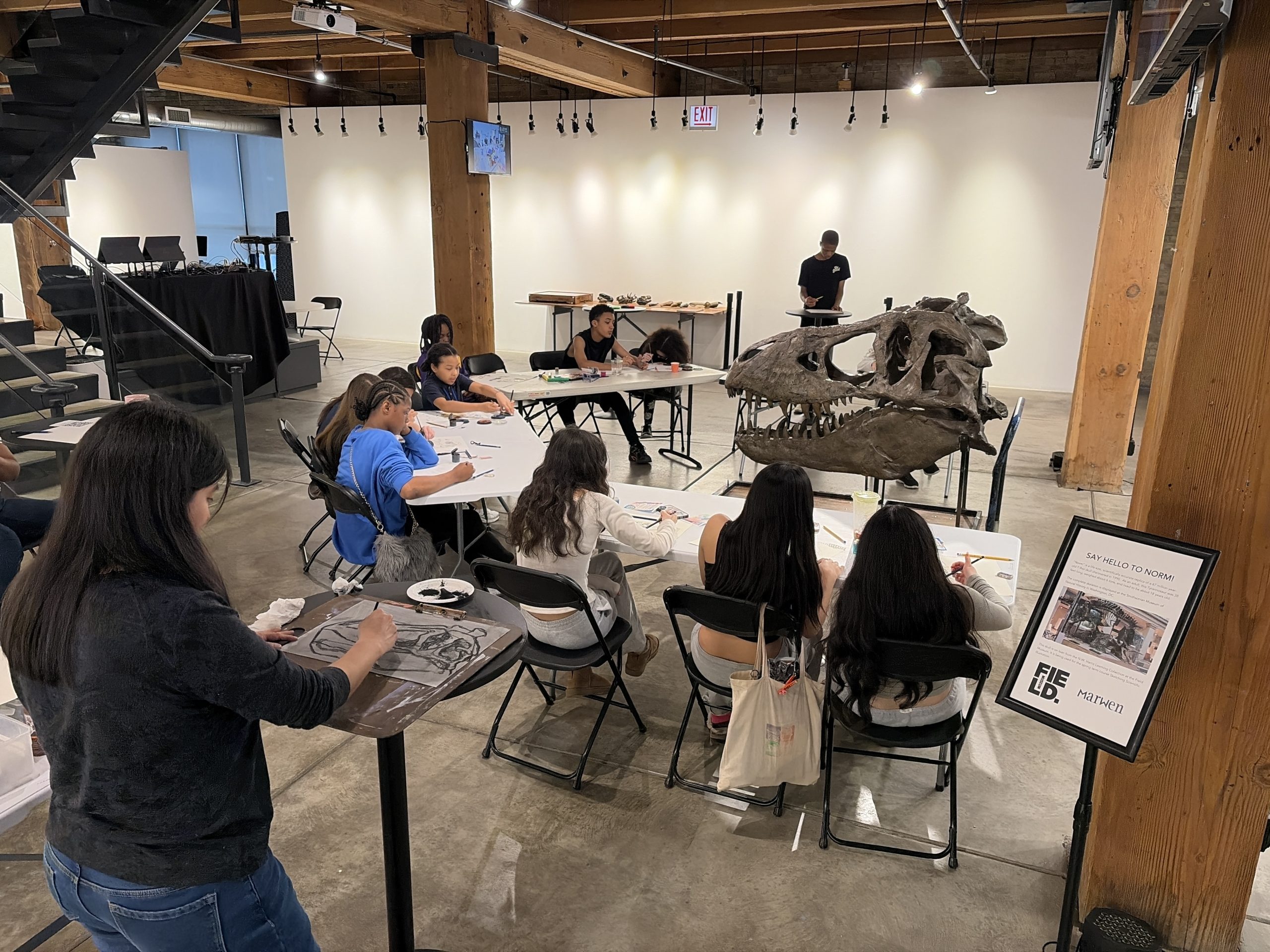 Group of students sketching at long tables in a museum studio, with a large dinosaur skull model on display nearby.