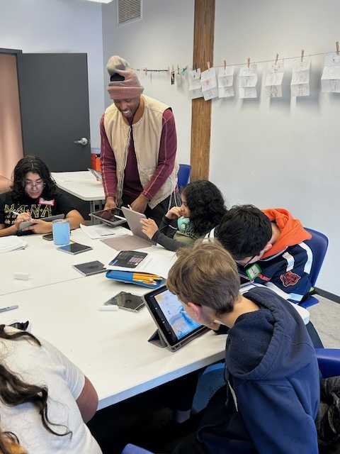 Teacher assists students using tablets at a classroom table, with papers on a clothesline in the background.