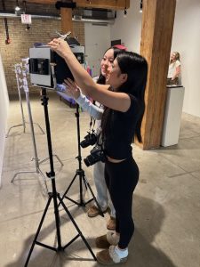 Two women in a studio adjust a tablet mounted on a tripod for a photo shoot, with cameras around their necks nearby.