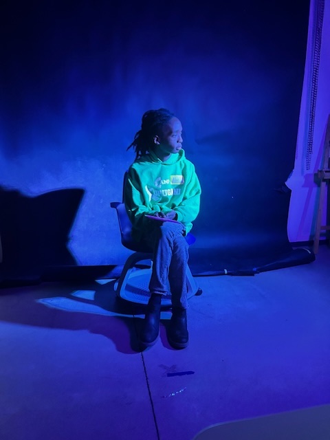 Person sitting on a chair in a blue-lit studio, wearing a bright green hoodie and jeans, looking off to the side.